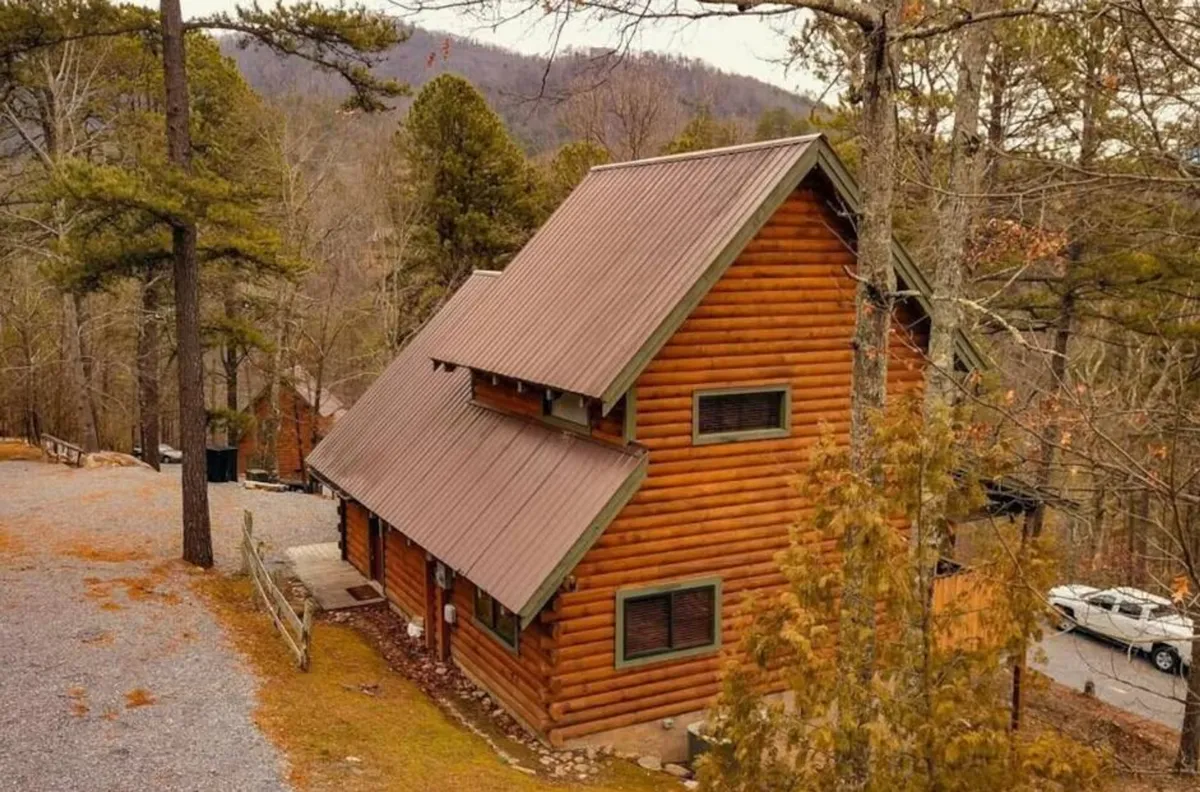 A cozy log cabin with a metal roof nestled among mature trees. The front porch offers a perfect spot for sunset watching, wildlife spotting, or simply unwinding in nature. The scene radiates warmth and tranquility, hinting at the exclusivity of the location.