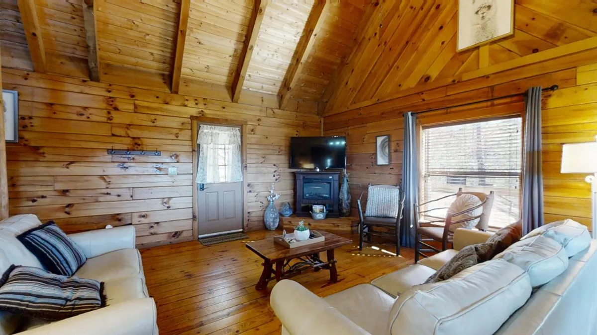 A comfortable seating area with a sofa, coffee table, television, and a window dressed with curtains. Wooden walls create a rustic ambiance, while the setup suggests a spot for entertainment and togetherness.