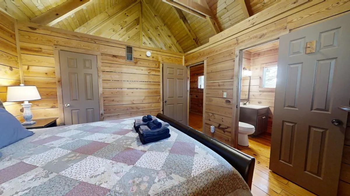 This image features a spacious king bedroom with a cozy quilted bed, a lamp on the bedside table, warm wooden walls, and an open bathroom door visible in the background. Soft natural light filters into the space, contributing to a tranquil, homey atmosphere.