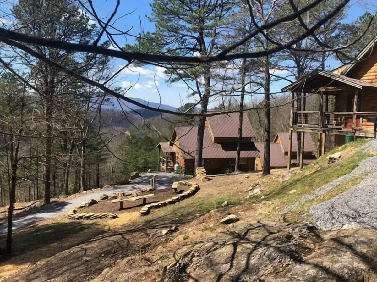 An inviting exterior view of wooden cabins flanked by trees, with a stone pathway leading to a visible fire pit area. The background reveals beautiful mountain vistas, suggesting opportunities for gathering and enjoying the outdoors.