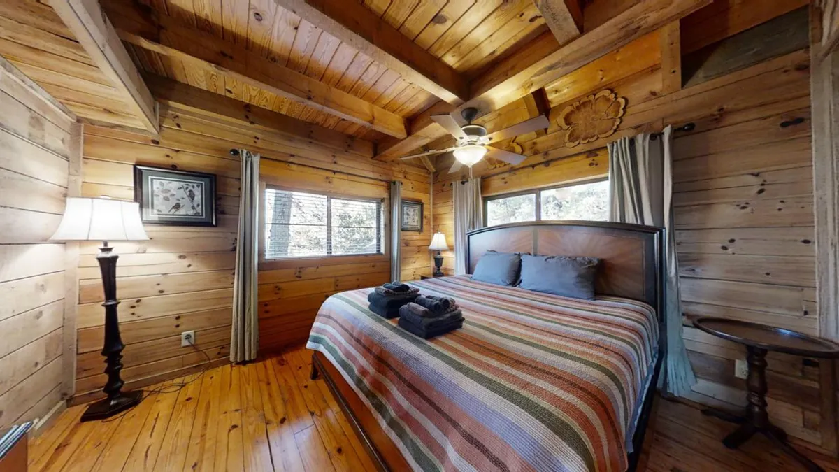 A comfortable bedroom with a striped blanket on the bed, charming wooden ceiling and walls, soft lighting from bedside lamps, and windows letting in natural light. The space feels airy and serene, ideal for relaxation.