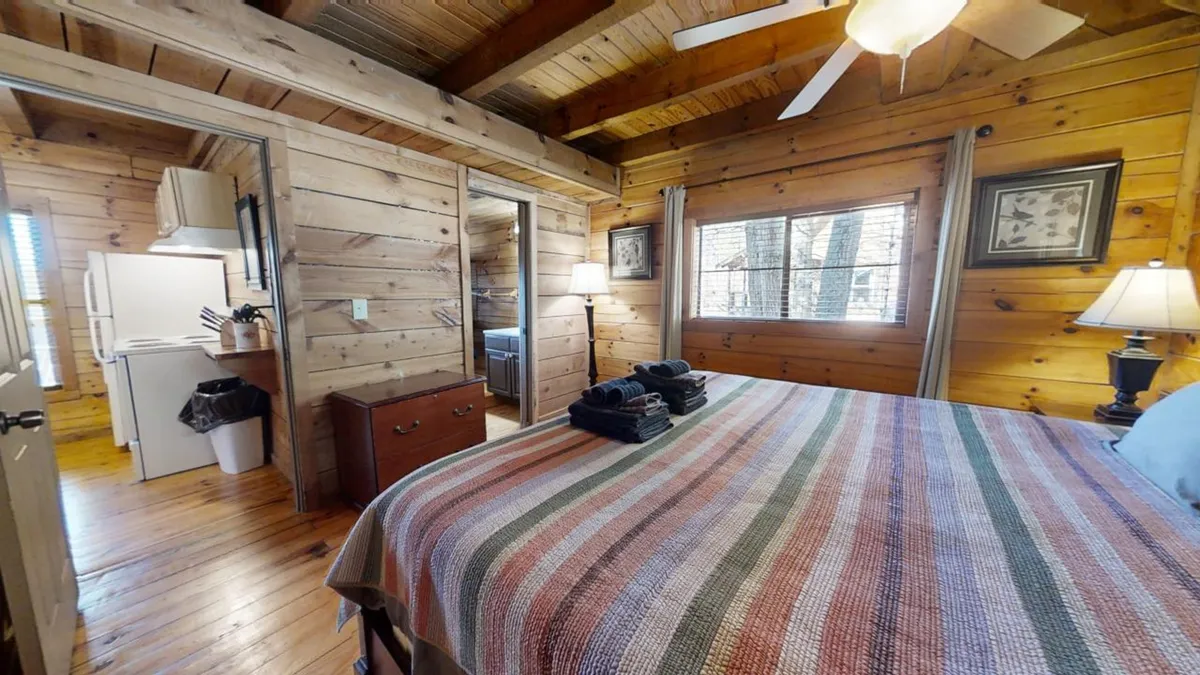 A cozy bedroom with a bed covered in a striped blanket sits against natural wooden walls. Soft lamplight and framed artwork add a warm, inviting atmosphere, while a kitchenette area hints at self-sufficiency for intimate retreats. The window offers a hint of the natural surroundings, connecting the interior to the outdoors.