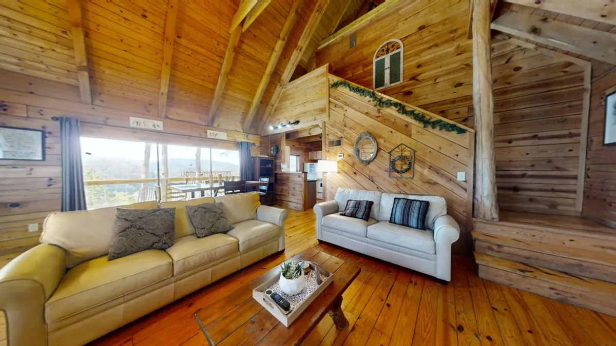 A warm and welcoming living room featuring plush sofas, a prominent wooden staircase, large windows offering plenty of natural light, and a central coffee table. The environment feels inviting—perfect for socializing, relaxing, and enjoying the view.